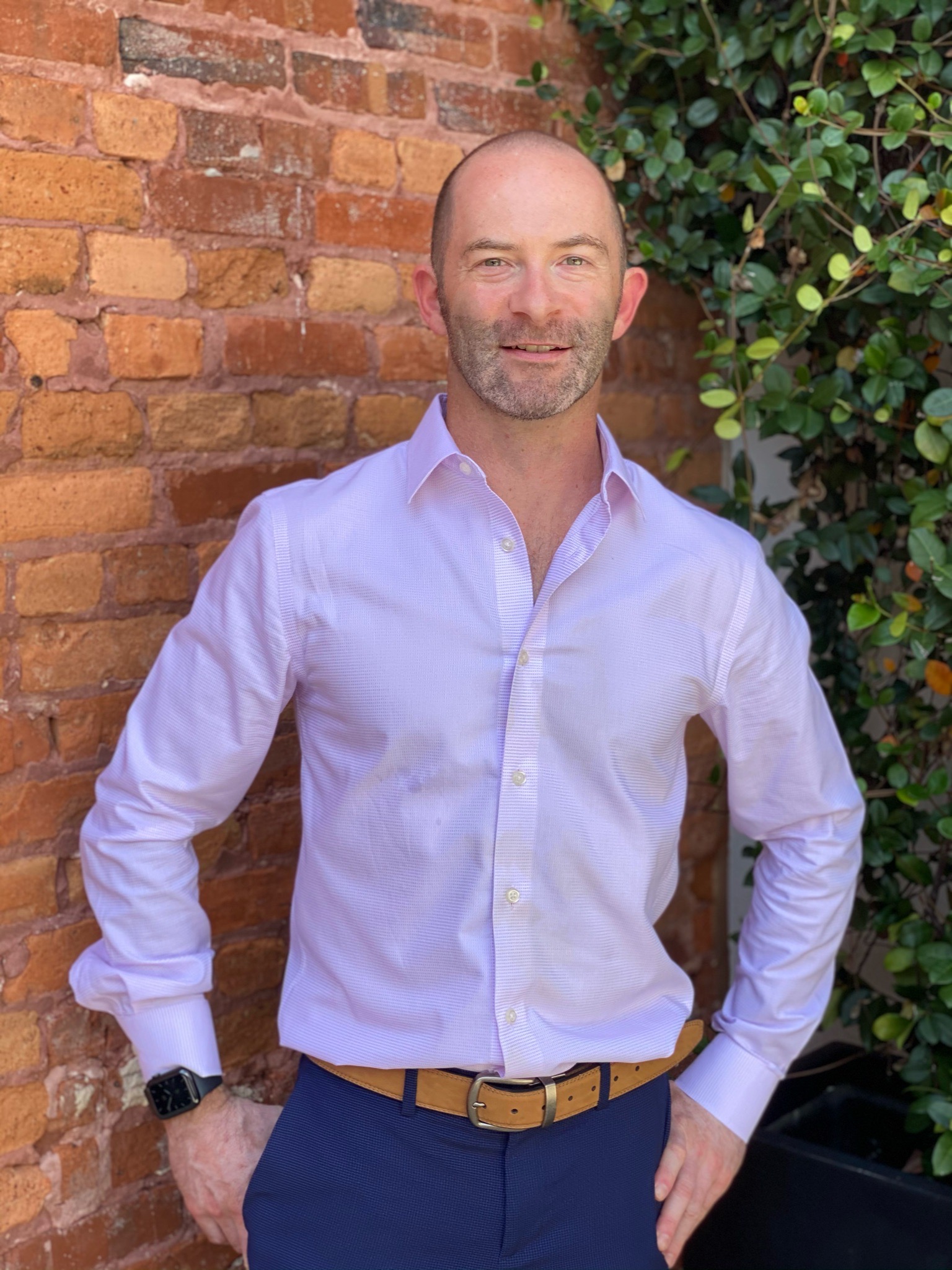 Geoffrey Fahey pink shirt and blue pants standing in front of a brick wall and plant